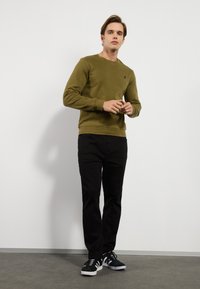 Sudadera verde oliva, con cuello redondo, puños y dobladillo de punto, combinada con pantalones negros. Unos zapatos deportivos en blanco y negro complementan el conjunto.