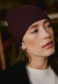 Burgundy ribbed knit beanie with a close-fitting design, paired with a cream sweater. Subtle shine on lips and small hoop earring visible.