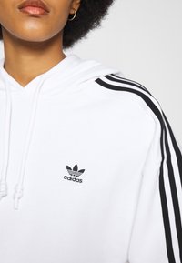 Hoodie blanc avec logo Adidas noir et trois bandes noires le long des manches. Équipé de cordons de serrage sur la capuche. Texture de tissu douce.