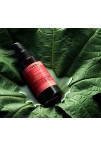 L'huile capillaire multi-usage Aveda Nutri Plenish dans un flacon sombre avec une pompe noire, étiquetée avec des accents rouges et roses, placée sur un fond de feuille verte.