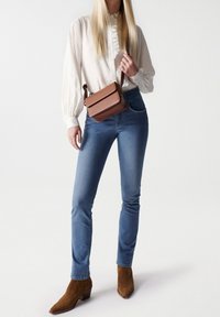 Hnědá crossbody kabelka s klopou, nošená přes bílou volánkovou blůzu, v kombinaci s modrými denimovými džíny a hnědými kotníkovými botami.