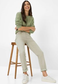 Donna sorridente seduta su uno sgabello di legno, indossando un maglione verde, pantaloni beige risvolti e sneakers bianche su uno sfondo bianco.