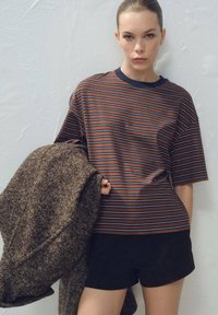 Pruhované hnedo-modré tričko s krátkym rukávom, doplnené čiernymi šortkami a držiace textúrovaný hnedý kabát.