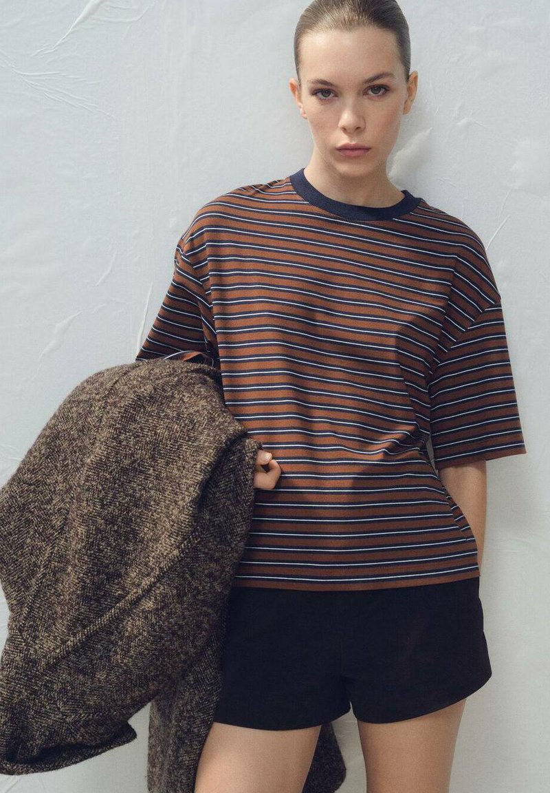 Pruhované hnedo-modré tričko s krátkym rukávom, doplnené čiernymi šortkami a držiace textúrovaný hnedý kabát.