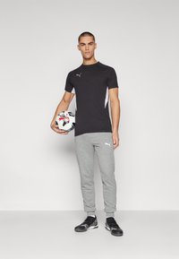 Cămașă sport neagră cu accente gri, asortată cu pantaloni de trening gri. Ținând în mână o minge de fotbal alb-negru. Pantofi sport negri.