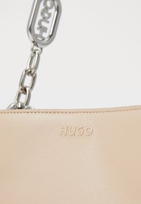 Sac en cuir beige avec un logo "HUGO" embossé, doté d'une poignée en chaîne argentée et d'une texture lisse. Design minimaliste avec des lignes épurées.