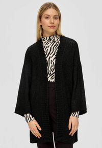 Schwarze gestrickte Strickjacke mit offenem Design, weiten Ärmeln, strukturiertem Rippenmuster, über einem schwarz-weißen gestreiften Oberteil getragen.