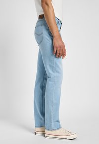 Muž stojící bokem, oblečený ve světlých modrých džínách s rovnými nohavicemi, bílém tričku a béžových plátěných teniskách na jednobarevném pozadí.