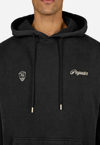 Čierna mikina s kapucňou a predným vreckom, so zlatým vyšívaním nápisu "Pegador" a logom na ľavej strane hrude.