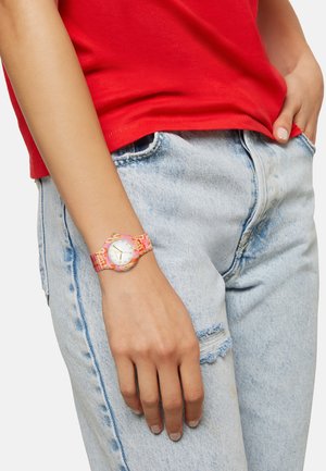 Farverig ur med et mønstret silikonearmbånd, rund guldfarvet kasse og hvid urskive. Båret på en hånd imod lyseblå slidte jeans.