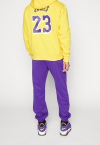 En person som bär en gul hoodie med "JAMES 23" i lila och vitt, lila svettbyxor och lila, rosa och vita sneakers, står mot en vit bakgrund.