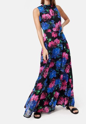 Femme portant une robe longue noire sans manches avec un grand imprimé floral rose et bleu, associée à des sandales noires ouvertes à l'avant, main sur la hanche.