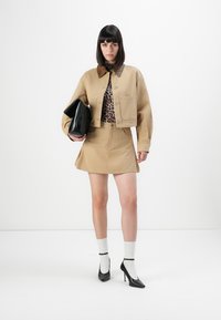 Kvinne i beige jakke og skjørt, leopardmønstret topp, hvite sokker med svarte high heels, som holder en stor svart clutchveske.