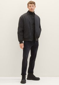 Schwarze Bomberjacke mit glatter Textur, Reißverschluss und gerippten Bündchen. Kombiniert mit dunklen Jeans und schwarzen Schnürstiefeln.