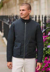 Navy blue bomber jacket with a high collar, front zip, and two buttoned pockets; worn over light-colored pants against a floral backdrop.