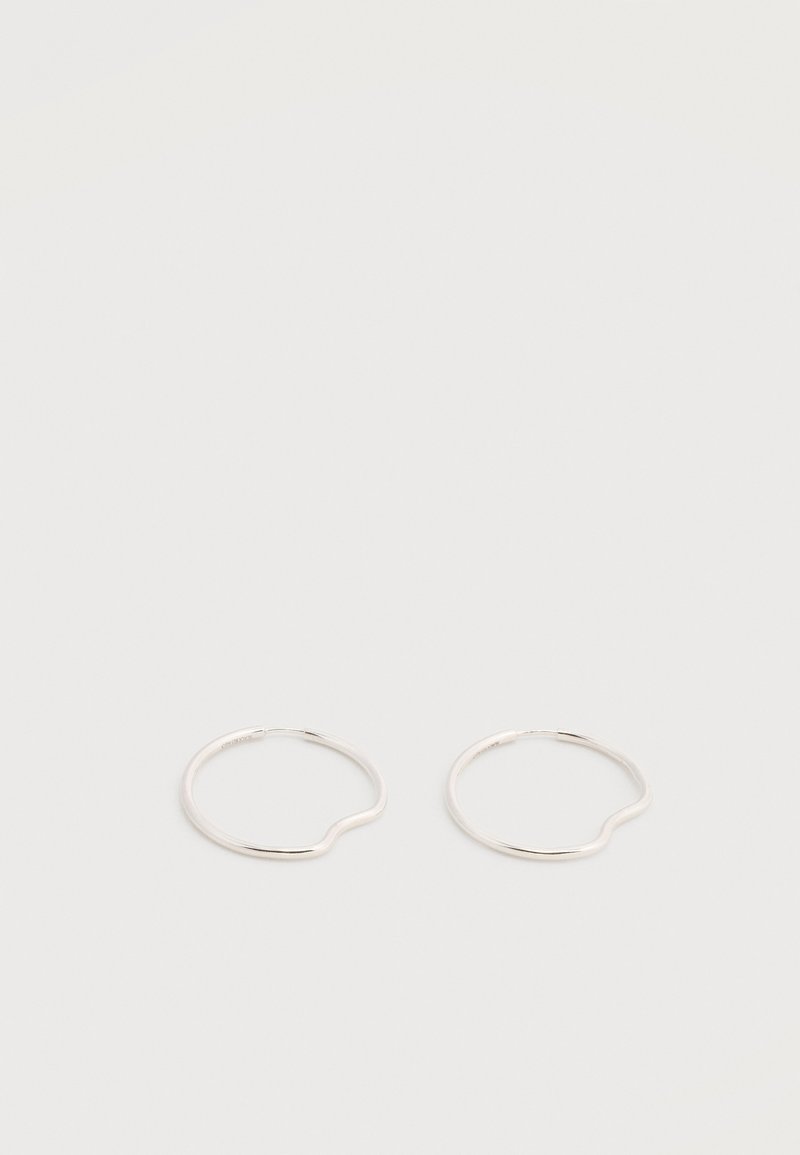 Δύο απλοί, λεπτοί ασημένιοι κρίκοι σκουλαρίκια με μια διακριτική καμπυλωτή λεπτομέρεια σε λευκό φόντο.