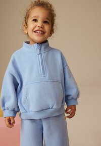 Sweatshirt en polaire bleu clair avec un col zippé mi-hauteur et une poche avant, associé à un pantalon de survêtement en polaire bleu clair assorti. Texture douce.