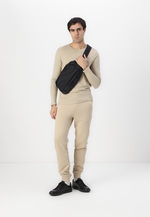 Cangurera negra con múltiples cremalleras, llevada cruzada por el cuerpo por un hombre que viste una camisa de manga larga beige y pantalones de chándal a juego. Zapatillas negras.
