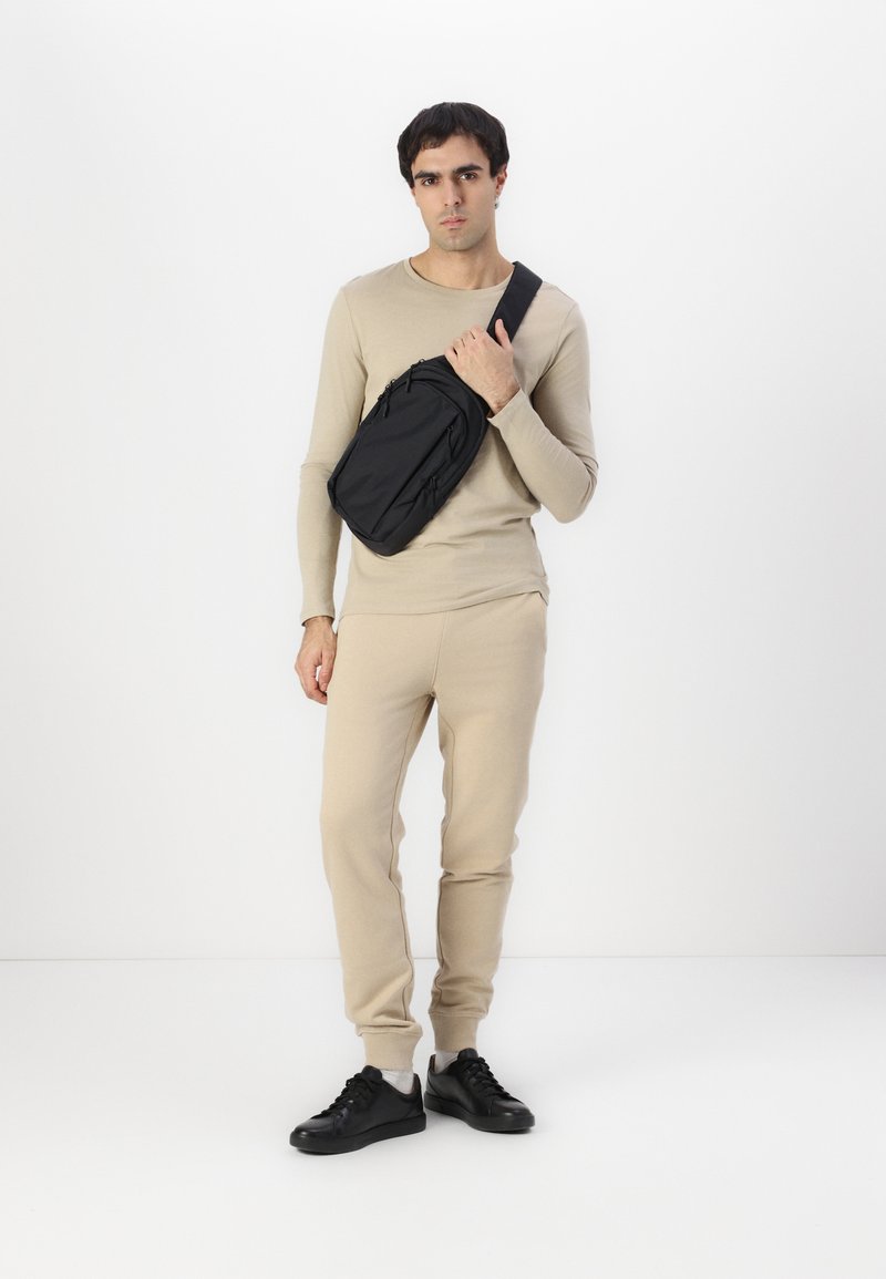 Cangurera negra con múltiples cremalleras, llevada cruzada por el cuerpo por un hombre que viste una camisa de manga larga beige y pantalones de chándal a juego. Zapatillas negras.