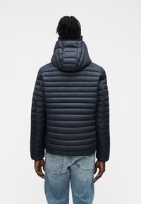 Tengerészkék steppelt mintázatú, kapucnival ellátott pufi dzseki. Bordázott mandzsettákkal és oldalsó varrásokkal rendelkezik, világoskék farmernadrággal párosítva.