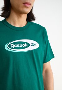 Žalias medvilninis marškinėliai su dideliu baltu ir šviesiai mėlynu ovaliu logotipu, kuriame yra užrašas "Reebok" ir dizaino elementas kairėje pusėje.