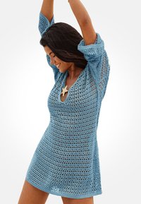 Mujer sonriendo con los brazos levantados, vistiendo un vestido de punto abierto azul claro sobre un traje de baño, con un accesorio de estrella de mar.