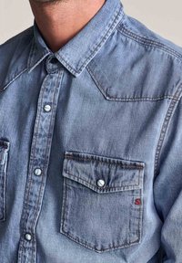 Camicia di jeans azzurro chiaro con colletto classico, chiusura frontale con bottoni e due tasche sul petto con bottoni a pressione. Caratteristiche cuciture a contrasto.