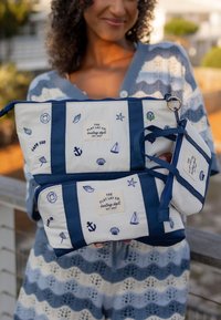 Femme tenant trois sacs bleu et blanc à thème nautique étiquetés "The Flat Lay Co. boating dept. Est. 2017" en extérieur sous une douce lumière du jour.
