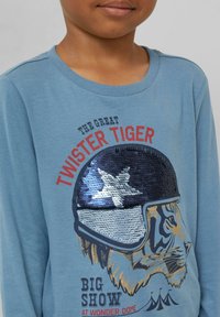 Blaues Langarmshirt mit einem Tiger-Graphic, der einen Paillettenhelm trägt, mit dem Text "DER GROSSE TWISTER TIGER" und bunten Akzenten. Aus Baumwollmaterial.