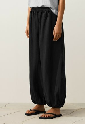 Personne portant un pantalon élastique noir ample aux chevilles, un haut gris et des sandales noires en tongs, debout sur un sol carrelé.