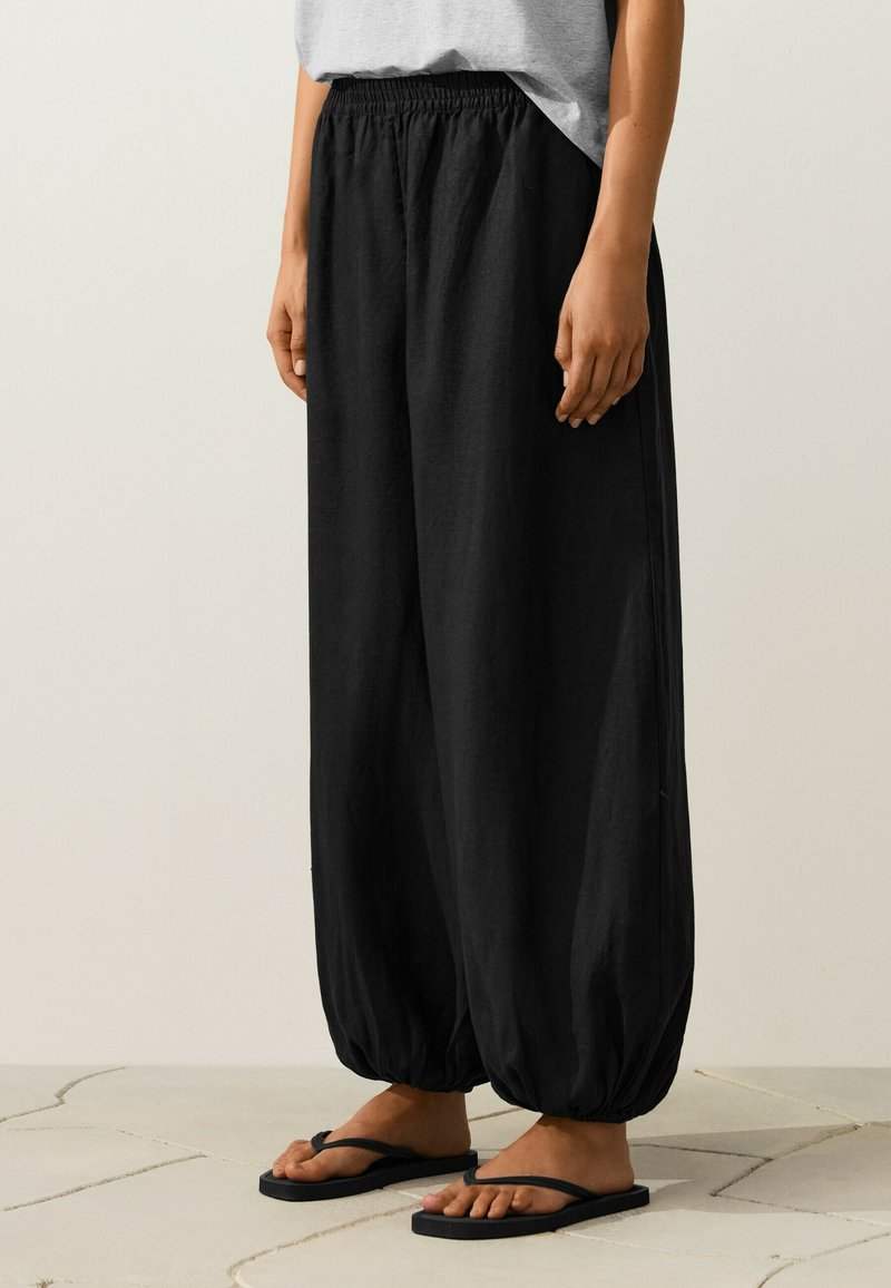 Personne portant un pantalon élastique noir ample aux chevilles, un haut gris et des sandales noires en tongs, debout sur un sol carrelé.