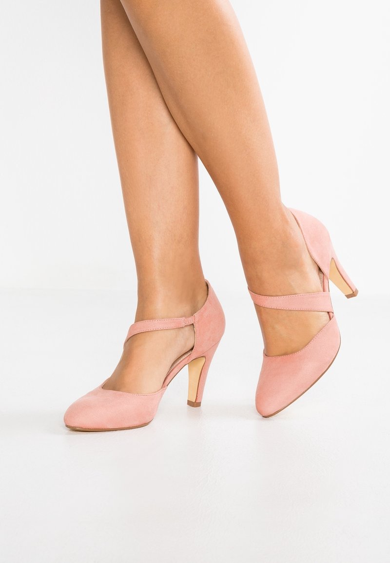 Jambes portant des escarpins Mary Jane en daim rose avec une bride sur le dessus du pied, debout sur un sol et un fond blancs.