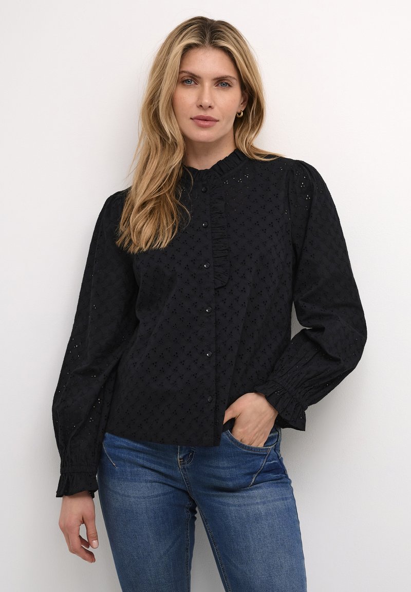 Schwarze Bluse mit Rüschkragen, langen Puffärmeln und strukturiertem Muster. Kombiniert mit hellblauen Jeans.