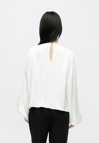 Blouse blanche à manches longues avec un détail goutte d'eau dans le dos, fabriquée en tissu lisse ; coupe légèrement ample avec des manches froncées, associée à un pantalon noir.