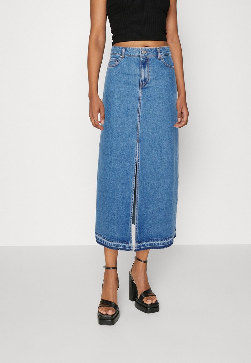 Femme portant une jupe midi en denim bleu taille haute avec une fente à l'avant, un top court noir et des sandales noires à talons plateformes.