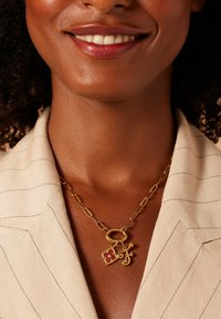 Gouden ketting met een ronde schakel en twee bedels: een hart met een rode accent en een gestileerde letter "F." Draagt over een gestreepte beige top.