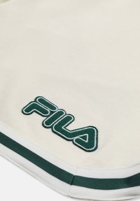Tissu blanc avec un patch logo FILA vert foncé et un détail de bordure rayée assortie vert et blanc.