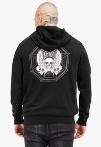 Sort hættetrøje med et stort grafisk print på ryggen, der viser kranier, vinger og ordet "TAPOUT" i fede bogstaver, med en struktureret finish.