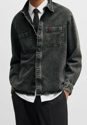 Mann trägt schwarze, gewaschene Jeansjacke über weißem Hemd und schwarzer Krawatte, mit Händen in den schwarzen Hosentaschen, vor einem schlichten Hintergrund.