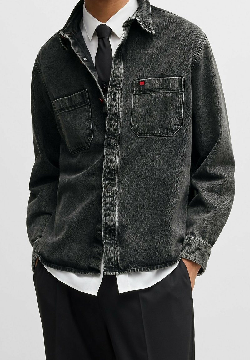 Homme portant une veste en denim noir délavé par-dessus une chemise blanche et une cravate noire, les mains dans les poches d'un pantalon noir, sur fond uni.