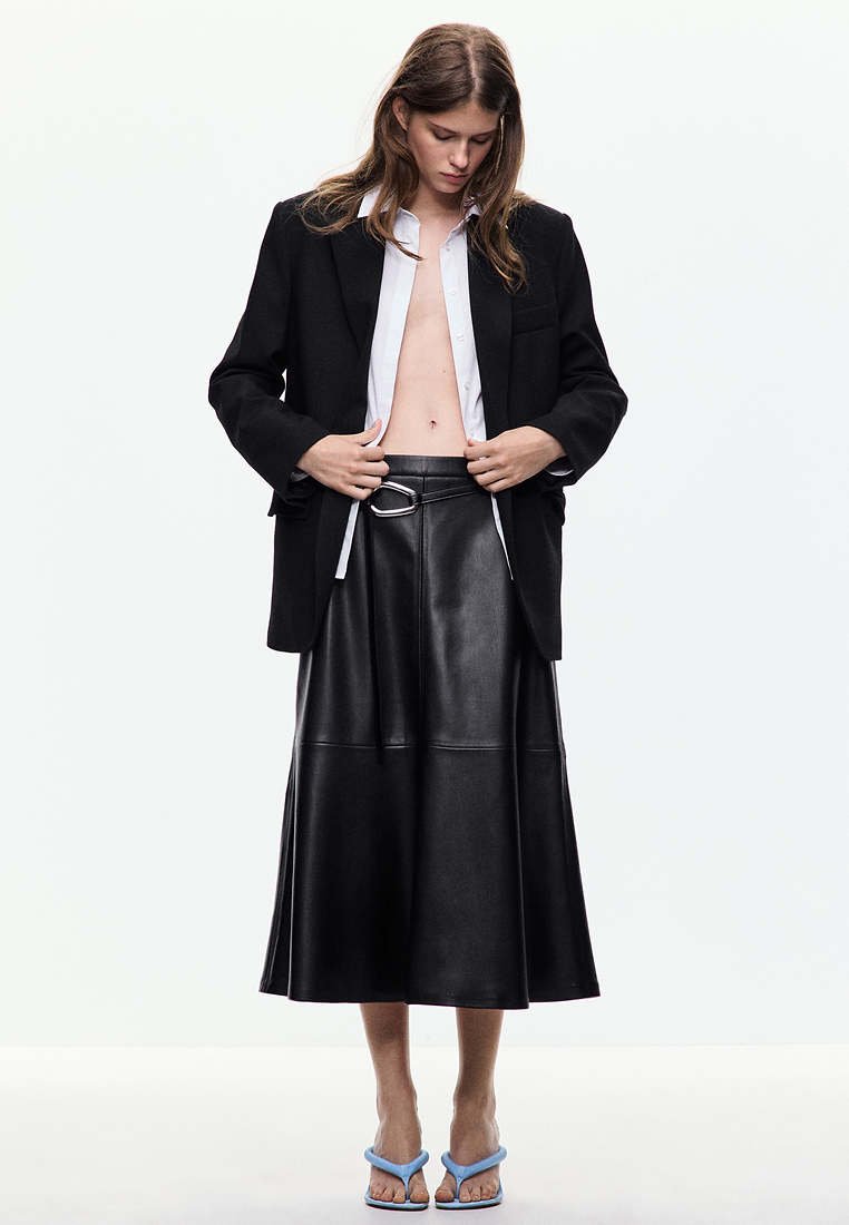 Femme portant un blazer noir par-dessus une chemise blanche ouverte, une jupe midi en cuir noire avec ceinture, et des sandales bleues à talons, debout devant un fond uni.
