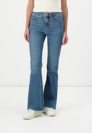 Flared blue jeans in denim, featuring a mid-rise waist, copper button accents, and a raw hem. Paired with white sneakers.