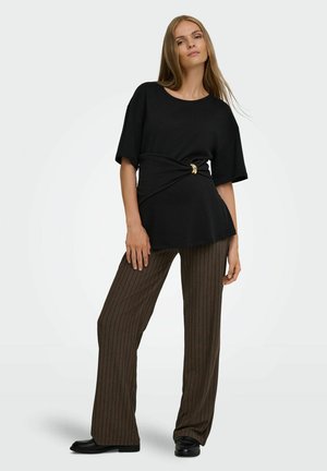 Mujer con una camiseta negra de manga corta con detalle de anillo dorado y pantalones anchos marrones con rayas y zapatos negros.