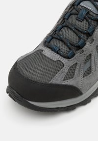 Grå tursko med mesh- og semsket skinnmateriale, teksturert overflate, svart gummitåhette, og blå detaljer på snøringene.