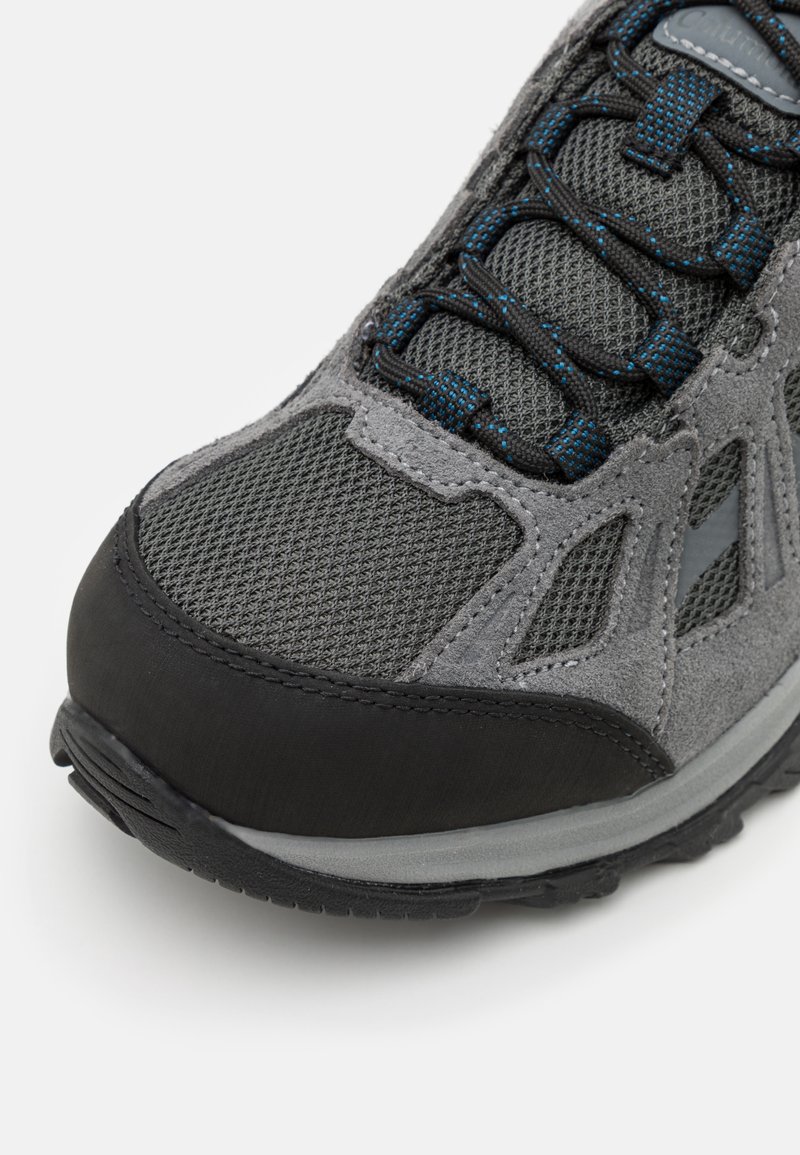 Grijze wandelschoen van mesh en suede materialen, met een gestructureerd oppervlak, zwarte rubberen neus en blauwe accenten op de veters.