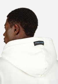 Weißer Hoodie mit einer großen, weichen Kapuze. Ein schwarzes Etikett im Nackenbereich trägt die Aufschrift "PEGADOR." Der Stoff hat eine strukturierte Oberfläche.