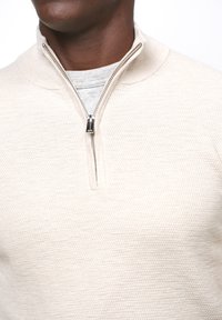 Maglione beige chiaro a metà zip in maglia testurizzata, con zip in metallo e colletto curvo sopra uno strato interno grigio.
