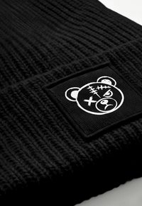 Black knitted beanie featuring a textured pattern and a square patch with a white graphic of a bear with stitches and an expressive face.