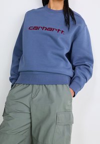 Sudadera azul con cuello y puños acanalados, con el logo "carhartt" bordado en color burdeos en el pecho. Combinada con pantalones cargo verde claro.