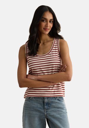 Mujer de cabello largo y oscuro, vistiendo un top sin mangas a rayas rojas y blancas y jeans azules, de pie con los brazos cruzados sobre un fondo blanco.